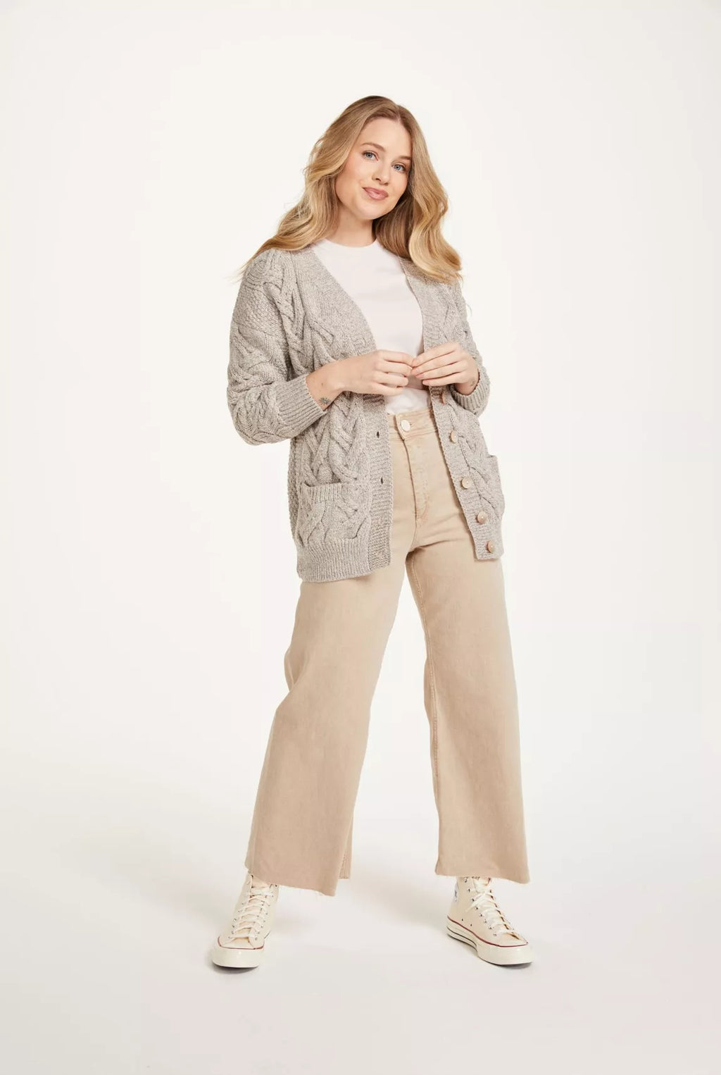 Vooraanzicht van een vrouw, zij draagt een beige gebreid vest, wit shirt, beige wijde jeans en beige sneakers. 