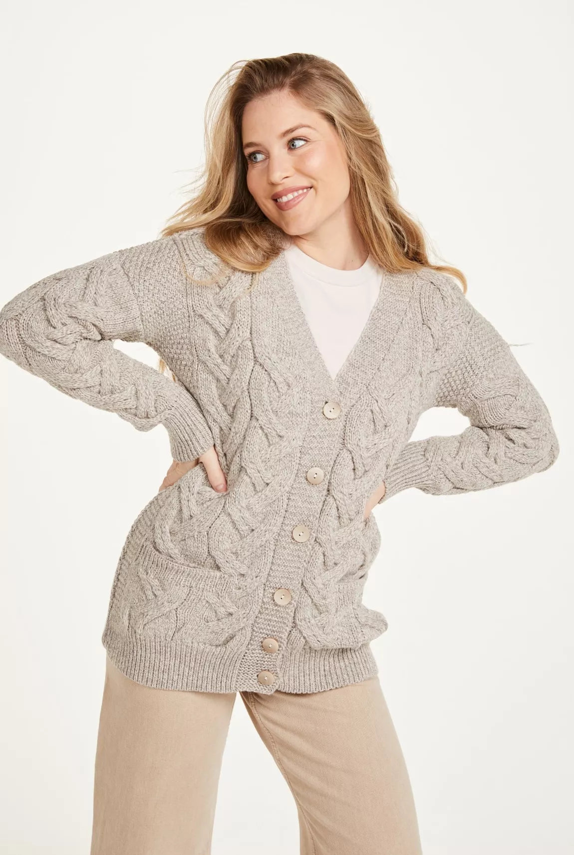 Vrouw draagt beige kabelgebreid vest. Vest valt over de heupen, heeft een knoopsluiting van 6  knopen en twee zakken.