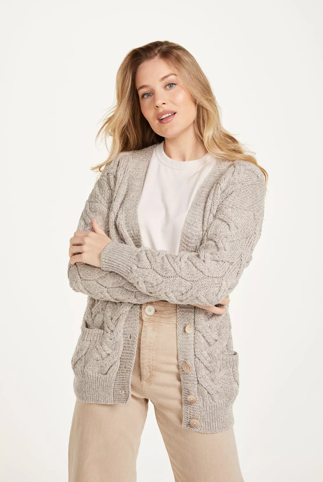 Vrouw draagt een openvallend vest. Het bovenstuk is kabelgebreid, beige en heeft 6 knopen. De schoudernaden zijn verlaagd en rondom afgewerkt met een breed boord.