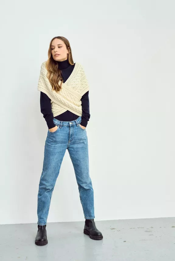 Vrouw draagt een crèmekleurige gebreide wikkel sjaal over een zwarte coltrui, gecombineerd met blauwe high-waisted jeans en zwarte enkellaarzen.