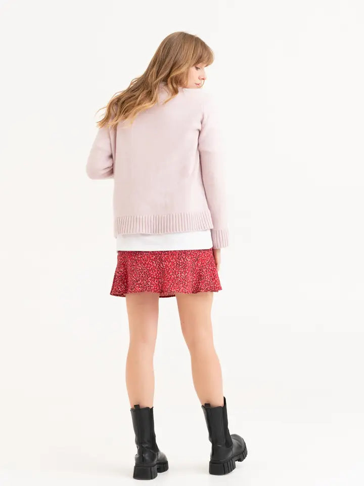 Van een roze gebreide trui wordt het achteraanzicht gepresenteerd. Het brede boord aan de zoom is goed zichtbaar. Gecombineerd met een wit shirt, rood rokje en zwarte laarzen.