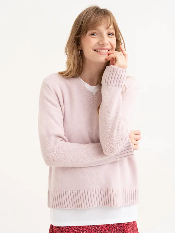 Model draagt een gebreide, zacht roze trui met een V-hals en brede boorden. het recht vallende silhouette wordt gecombineerd met een wit shirt en een rood rokje met een abstracte print.