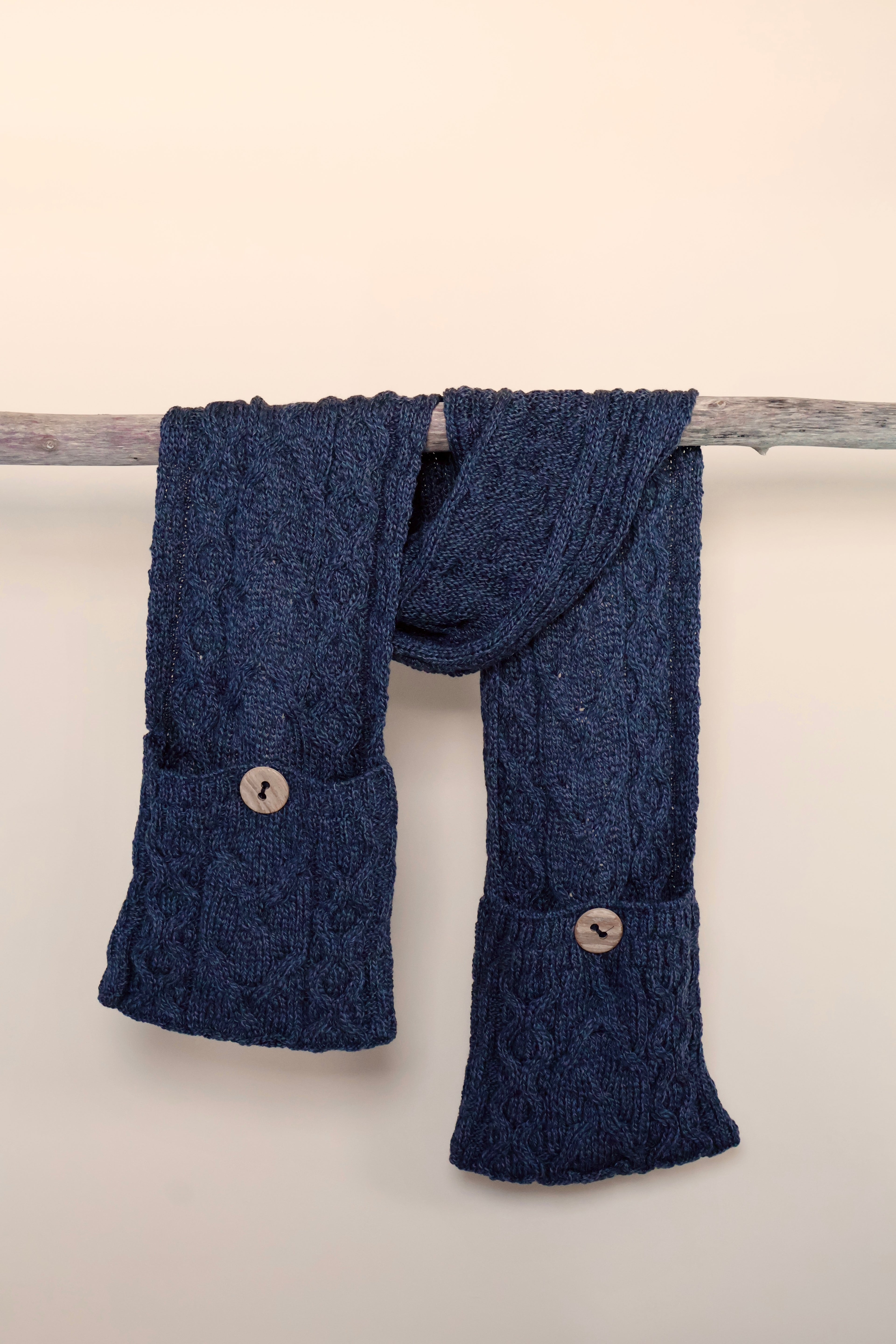Sjaal gedrapeerd over een houten tak. Het blauwe breisel heeft zakken met knopen. 