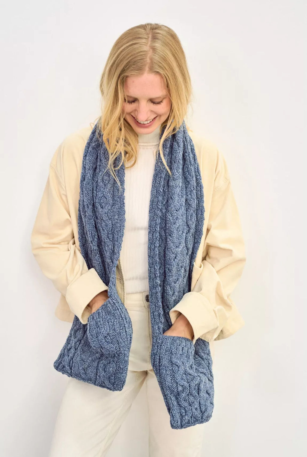 Vrouw draagt een witte outfit met een grote blauwe sjaal met zakken. Haar handen accentueren dit praktische detail.