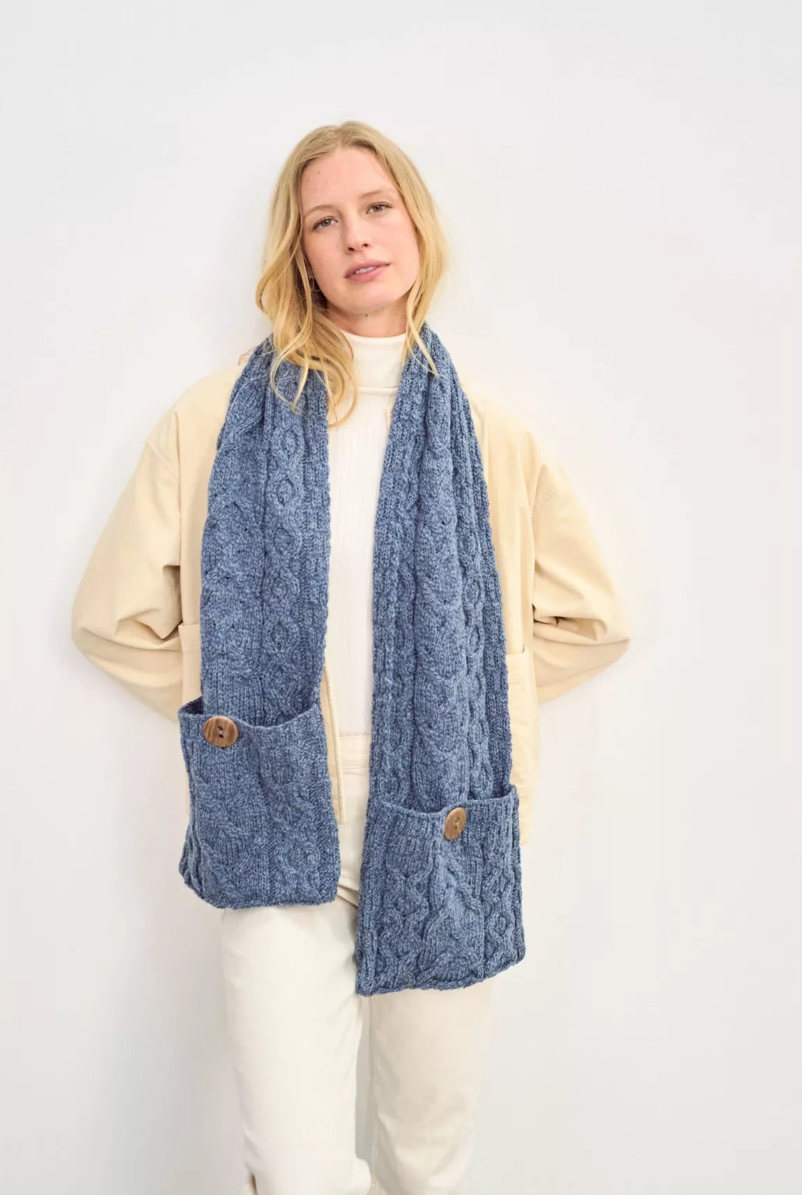 Een kabelgebreide sjaal in een jeans blauwe kleur wordt gedragen door een vrouw. De focus ligt op de door een knoop gesloten zak op de uiteinden van deze winter accessoire.