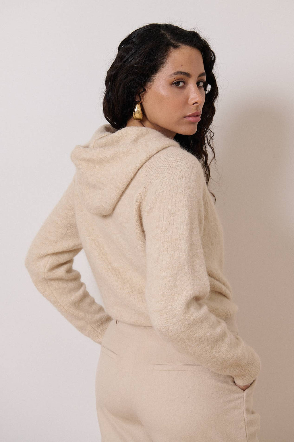 Een achteraanzicht van een vrouw met een beige outfit. De nadruk ligt op de fijn gebreide trui met een capuchon.