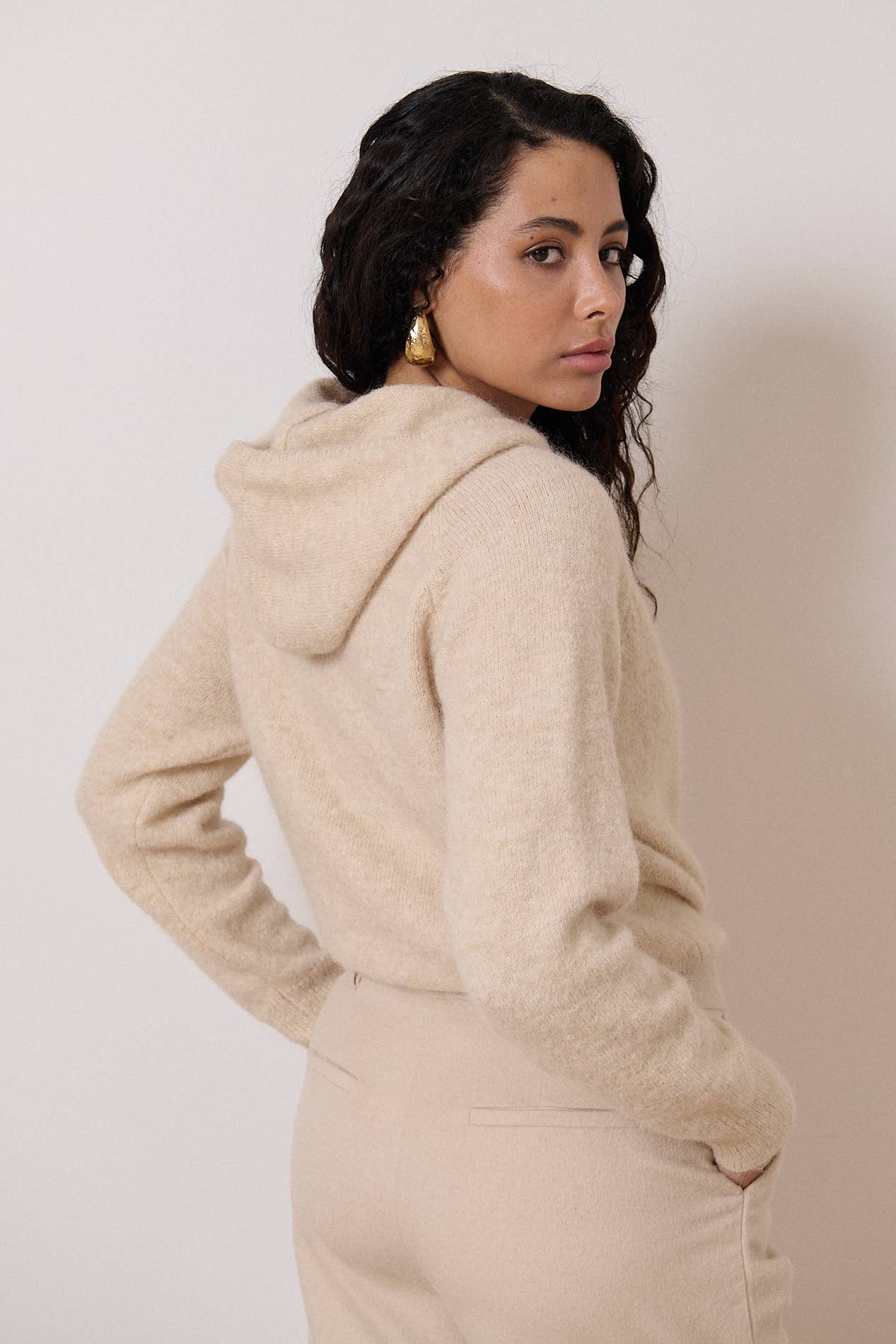Een achteraanzicht van een vrouw met een beige outfit. De nadruk ligt op de fijn gebreide trui met een capuchon.