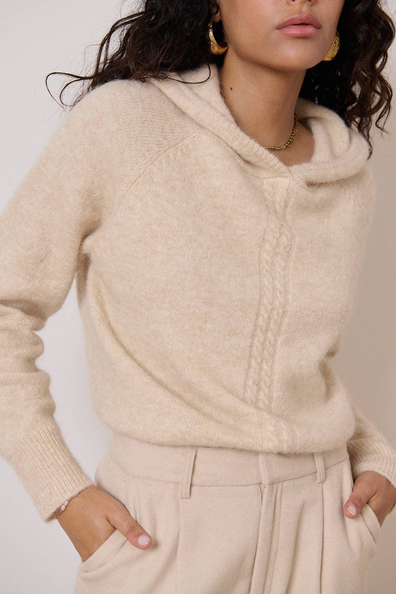 Een vrouw met een beige trui en broek aan. De capuchon geeft het model extra comfort. Het voorpand is verfraaid met een kabel detail.