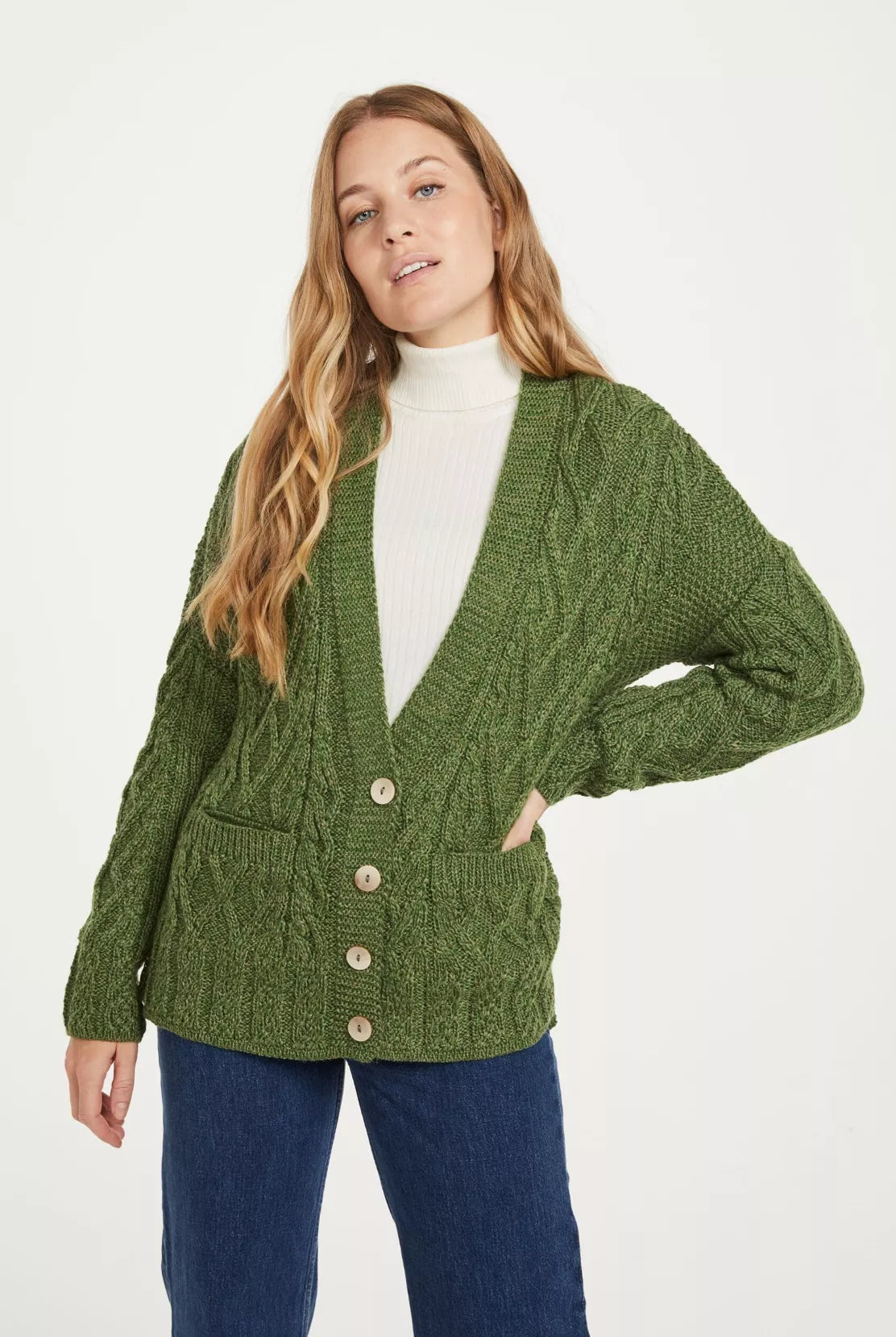 Vrouw draagt een groen kabelgebreid vest met vier knopen, twee zakken en V-hals. Het kledingstuk wordt gecombineerd met een witte coltrui en jeans.