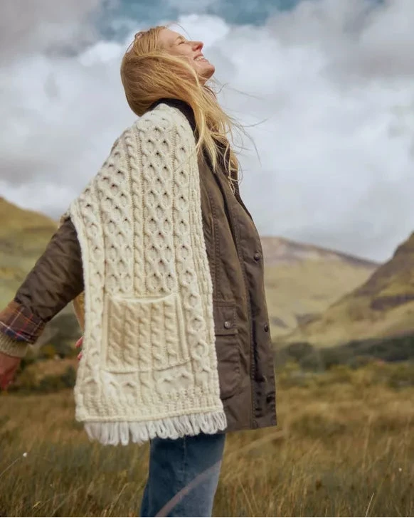 Vrouw staat in de natuur. Ze draagt een bruine jas met daar overheen een witte grof gebreide sjaal met zak en franjes.