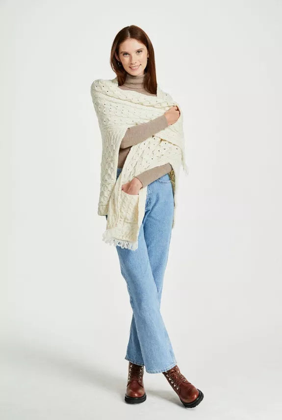 Vrouw draagt een crèmekleurige gebreide omslagsjaal met kabelpatroon en franjes, over een beige coltrui en lichte jeans, met bruine veterlaarzen.