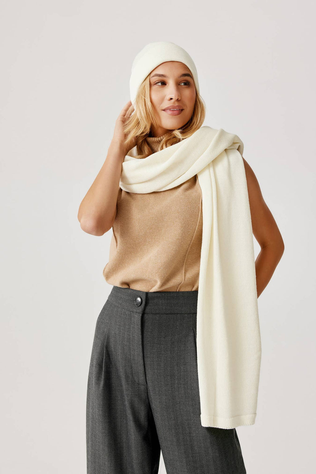 Vrouw draagt een grijze pantalon en beige top. Ze heeft een witte muts en sjaal omgeslagen.